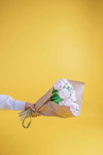 A person extends their arm holding a bouquet of fresh, pale pink tulips with green stems, wrapped in light brown craft paper and tied with a green ribbon, against a vibrant yellow background. The tuli