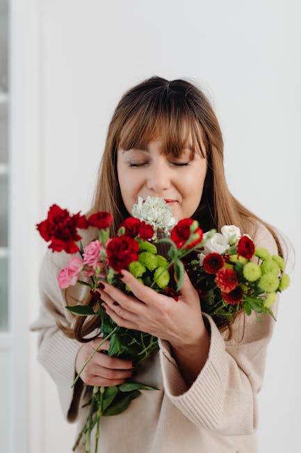 A woman with light brown hair and bangs, dressed in a beige sweater, is holding a vibrant bouquet of fresh flowers close to her face with her eyes closed and a gentle smile. The bouquet features a mix