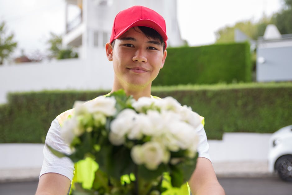 A delivery person wearing a red cap and a high-visibility vest is holding out a floral gift of white roses and other white blooms wrapped in clear cellophane and tied with a green ribbon, with a subur