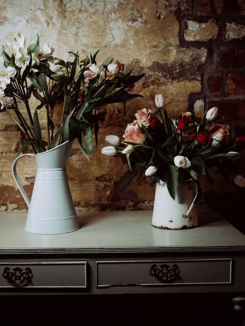 A photograph showcasing two floral arrangements in vintage-style metal jugs placed on a grey sideboard. The left jug contains white lilies with long green stems and leaves, arranged to display their o