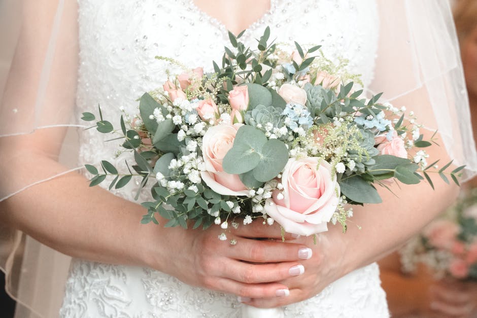 A bride holds a romantic floral bouquet featuring blush pink roses, white spray roses, and green eucalyptus leaves with silver-green tones, arranged in a loose, natural style. The bouquet includes del