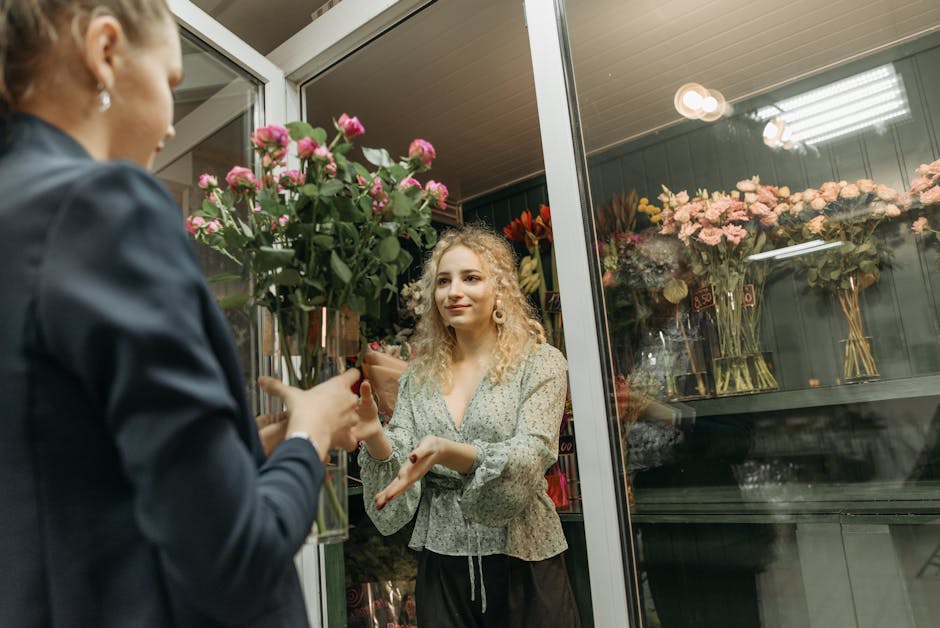 A customer in a dark jacket hands a bouquet of pink roses and green foliage to a young woman with curly blonde hair, wearing a light-colored floral blouse, inside a flower shop with a glass door. Behi