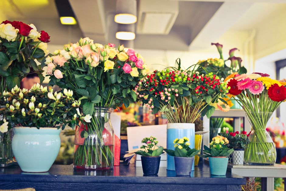 A display of fresh floral arrangements and bouquets on a dark wooden table inside a well-lit flower shop. The arrangement includes white, pink, and red roses in clear and pastel-colored vases, with vi
